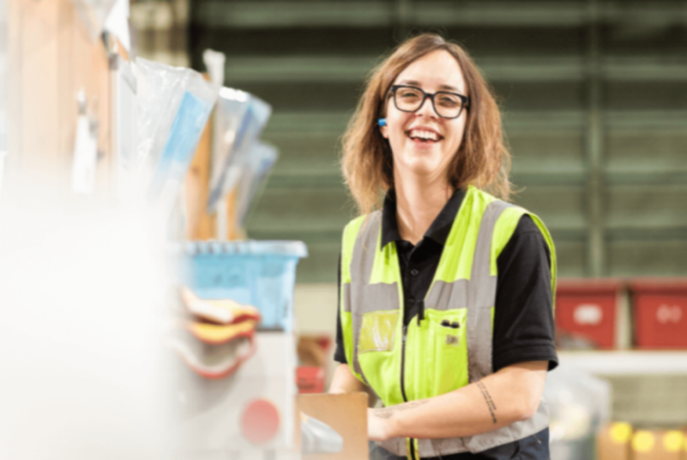 Smiling woman with brown hair wearing glasses and a high-visibility vest, standing in a warehouse environment with industrial shelving.